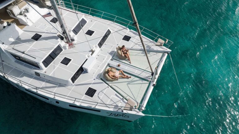 Large catamaran at anchor with charter guests relaxing on the bow trampolines. Charter Catamaran TAPAS Accommodates 10 guests in 5 cabins. All Inclusive week charters in the BVI starting at $25,000. Charter with Captain & Chef Onboard.